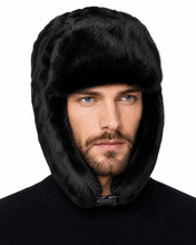 Load image into Gallery viewer, A close-up of a man wearing a black faux fur ushanka hood with ear flaps and a chin strap, paired with a black ribbed sweater, against a plain white background.