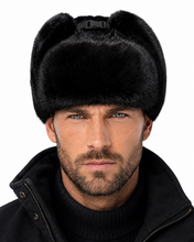 Load image into Gallery viewer, A close-up of a man wearing a black faux fur ushanka winter hat with a black buckle strap and a black zip-up jacket against a white background.