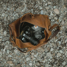 Load image into Gallery viewer, The Dagny Weekender | Large Leather Duffle Bag on gravel with camera inside, showcasing spaciousness and organization for travelers.