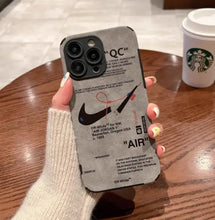 Load image into Gallery viewer, Luxury leather sneaker phone case featuring a sleek design and branding, held over a wooden table next to a coffee cup.