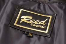 Load image into Gallery viewer, Close-up of Reed Est. 1950 label on genuine lambskin men's jacket, showcasing quality craftsmanship and classic design in black and gold.