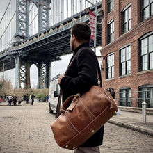 Load image into Gallery viewer, Man carrying The Dagny Weekender | Large Leather Duffle Bag near iconic bridge and brick buildings, showcasing style and durability.