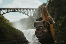 Load image into Gallery viewer, Woman exploring with The Hagen Backpack | Vintage Leather Backpack near a scenic waterfall and bridge.