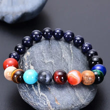 Load image into Gallery viewer, Eight Planets Natural Stone Bracelet with Blue Sand Stone, Red Agate, Tiger Eye, Lapis Lazuli, and Yellow Jade on a rock background.