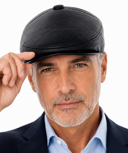 Load image into Gallery viewer, A close-up portrait of a man wearing a black leather flat cap and a navy blue suit jacket over a light blue dress shirt, adjusting the brim of the cap against a solid white background.
