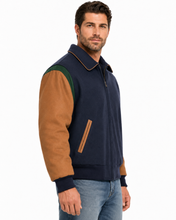 Load image into Gallery viewer, A man wearing a navy blue zip-up jacket with tan suede sleeves and green shoulder panels, paired with blue jeans, photographed against a plain white background.
