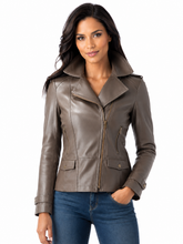 Load image into Gallery viewer, A woman modeling a taupe-brown real leather moto jacket with a stand-up collar and asymmetrical zipper, paired with blue jeans against a plain white background.
