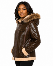 Charger l'image dans la galerie, A woman wearing a glossy brown real-leather hooded jacket with beige faux-fur trim on the hood, collar, cuffs, and hem, shown against a plain white background.
