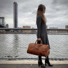 Load image into Gallery viewer, Woman standing with The Dagny Weekender | Large Leather Duffle Bag by a river in an urban setting
