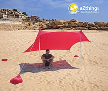 Load image into Gallery viewer, Person sitting under red beach shade canopy with sandbag anchors, designed by eZthings, on a sunny beach. Provides UV protection.
