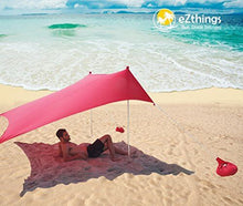 Load image into Gallery viewer, Man relaxing under eZthings UV sun shade beach shelter on sandy shore, sunny day, red canopy with sandbag anchors, ocean view.
